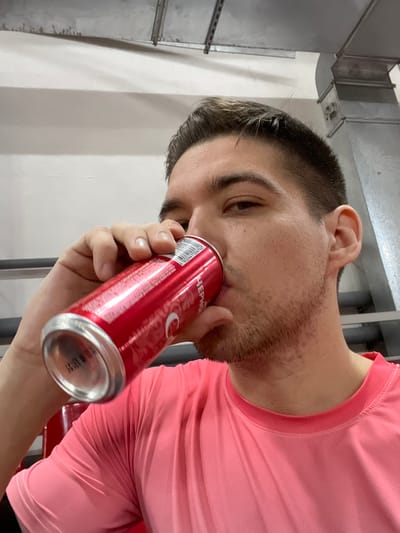 A man in a pink athletic shirt is tilting his head back and to the left, holding a red soda can near his nose. His close-cropped hair and stubble are visible. The can is prominently displayed, showing its label and the bottom with printed dates. Background elements include ventilation ducts and a white wall, suggesting an industrial or institutional setting. The photograph is taken from a low angle, emphasizing the upward tilt of the man's head.