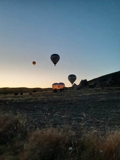 The image depicts a dawn scene in Cappadocia, near Avanos, Turkey, characterized by a clear, pale blue sky transitioning to a warm orange glow near the horizon. Several hot air balloons are airborne, with one prominent balloon in the mid-right displaying the "Butterfly Balloons" logo. To its left, two other balloons are illuminated from within by their burners, casting a warm orange and yellow light. Further left, a smaller balloon with orange and brown stripes is visible. The landscape below consists of rolling hills and a grassy, somewhat barren field with dry vegetation. In the background, distinctive rock formations, typical of Cappadocia's unique geology, rise from the terrain. The overall atmosphere is tranquil, suggesting an early morning hot air balloon tour.