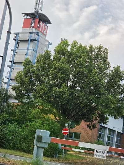 The image depicts the Rüsselsheim am Main fire station with its distinctive red and white warning tower, prominently displaying the number "112" for emergency services. A large, leafy tree stands in the foreground, partially obscuring the view of the building. A red and white striped barrier gate is lowered across a roadway in front of the fire station, indicating a closure or active response. A circular traffic sign with a red horizontal bar on a white background is also visible near the gate. The building itself features a brick facade and large windows. The sky is overcast with scattered clouds. The overall scene suggests a functional fire station environment, possibly during a drill or an active response.