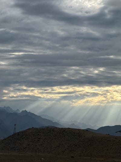 The image captures a dramatic mountain landscape in Chumkar, India, under a heavily clouded sky. The sun's rays pierce through breaks in the clouds, creating visible beams that illuminate the scene, particularly in the middle ground where they cast light upon a series of layered, silhouetted mountain ranges. The foreground features a dark, rocky hill, with a tall, slender metal structure, possibly a tower or part of a wind turbine, situated on its crest. A single figure, appearing red, is visible at the top of this structure. The terrain in the foreground is barren and dry, with sparse vegetation and several utility poles scattered across the landscape. The overall atmosphere is somber yet awe-inspiring, with the interplay of light and shadow highlighting the rugged beauty of the mountainous terrain.