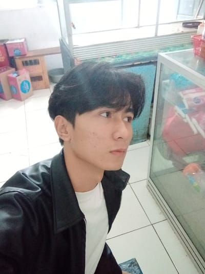A young male subject with dark, medium-length hair and a fair complexion is captured in a close-up indoor photograph, likely in Bojongherang, Indonesia. He wears a black collared jacket over a white t-shirt and looks towards the right of the frame with a neutral expression. The background displays a white tiled floor with grey grout lines. To the left, several pink cardboard boxes featuring blue circular logos are stacked near a wooden crate and a grey basin. To the right, an aluminum-framed glass display case contains various indistinct items, with a red plastic basket positioned on top. A light blue wall or surface is visible behind the display case, topped by white horizontal slats. The scene is illuminated by even, bright lighting.