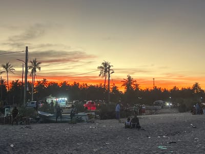 This image captures a sunset scene on a beach in Kalmunai, Sri Lanka. The sky displays vibrant shades of orange and yellow, with dark silhouettes of palm trees lining the horizon. Numerous people are present on the sandy beach, some standing in groups, others sitting, and some near small fishing boats. Several vehicles and a food stall with lights are visible in the background along the beachside. Debris litters the sand.