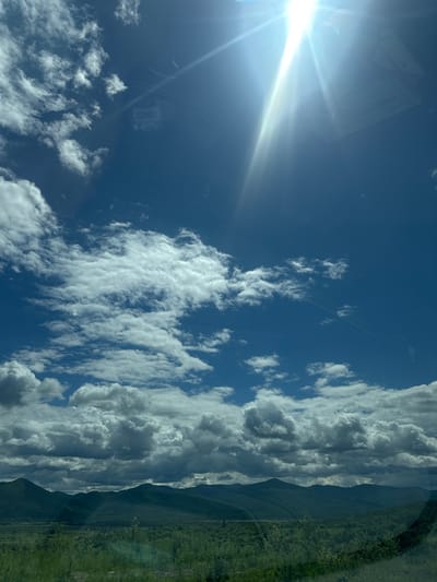 The image depicts a landscape scene under a bright, sunny sky with scattered white clouds. In the foreground, there is a grassy area and some trees. Beyond that, a range of green, tree-covered mountains stretches across the horizon. The sky is a vibrant blue, with the sun intensely bright in the upper right quadrant, casting sun flares. A faint reflection, possibly of a sticker or sign, is visible on what appears to be a window. The location is identified as Теплоозёрск, Russia. There are no people or animals visible in the frame.