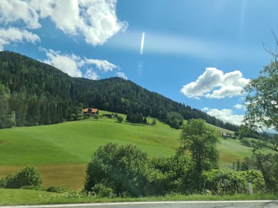 The image captures a picturesque rural landscape under a bright blue sky with scattered white clouds. A dense evergreen forest covers the slopes of a large hill that dominates the left side of the frame. Nestled on the hillside, amidst rolling green pastures, is a traditional alpine house with a red roof. The foreground shows a strip of road with a white line, bordered by lush green vegetation and trees. The scene is bathed in sunlight, creating a lens flare effect in the sky. The location is identified as Scheifling, Austria. There are no people or animals visible in the image.