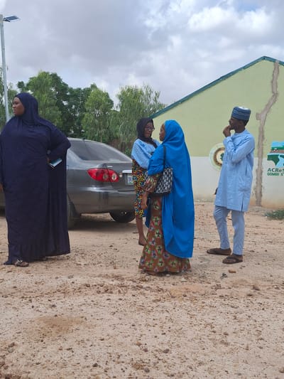 In Kundulun, Nigeria, under a cloudy sky, a group of four individuals stands near a light green building. On the left, a woman in a dark blue hijab and long dress stands with her back to the viewer, holding a notebook. To her right, two women in hijabs are conversing. One wears a blue hijab and a patterned dress, carrying a black handbag. The other, partially obscured, wears a blue garment and glasses. On the far right, a man in a light blue traditional outfit and a patterned cap stands, looking to his left with his hand on his chin. A grey Toyota car is parked behind the women, with its rear visible. The ground is a mix of sand and small stones. A street light with a solar panel is visible in the background. The building has a green trim along the roofline. A sign on the building reads "ACRes GOMBE ST".