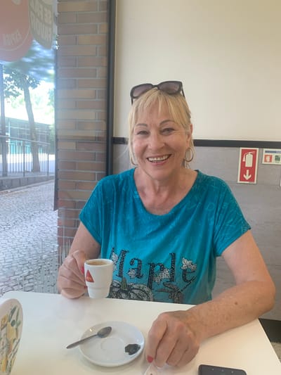 A middle-aged woman with blonde hair and sunglasses perched on her head sits at a white table indoors. She is wearing a turquoise t-shirt with a pattern of letters and designs. She is holding a small white espresso cup with the "DELTA" logo in her right hand, bringing it towards her mouth. In front of her is a white saucer with a spoon and a small dark blob, possibly dried coffee. The background shows a brick wall to the left and a plain wall with two red and white signs to the right. Through a window to the left, a cobblestone street and trees are visible, suggesting an outdoor setting beyond the building. The woman is smiling and looking at the camera. This scene appears to be in Odivelas, Portugal, likely at a cafe or similar establishment given the presence of coffee and the indoor seating arrangement.