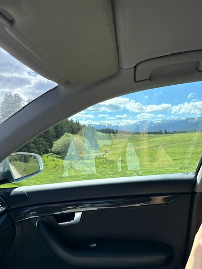 The image is taken from inside a car, looking out the passenger window and windshield. The interior of the car is visible in the foreground, including a portion of the door panel with a black leather finish, a chrome-colored door handle, and a speaker grille. Above the window, the car's headliner and part of the sun visor are visible.
Outside the car, a bright, sunny day is depicted. The view is of a lush green meadow with rolling hills. In the mid-ground, a dense forest of dark green trees is visible, likely pine or fir. Beyond the forest, a range of majestic, snow-capped mountains stretches across the horizon under a bright blue sky dotted with fluffy white clouds. There are reflections on the car window, suggesting the presence of a building or structure within the meadow, possibly a house or shed. The side mirror of the car is also visible, reflecting a dirt path and more green landscape.
The image does not provide any specific information or visual cues that would definitively place it in the city of Avramovo, Bulgaria.