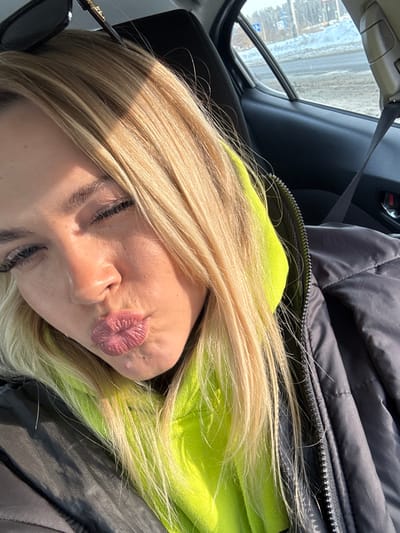 A close-up selfie taken inside a vehicle depicts a light-skinned female in L'vovskiy, Russia. The subject has long, straight blonde hair, with sunglasses perched atop her head. She is wearing a neon yellow hooded sweatshirt under a dark grey or black puffer jacket. Her eyes are partially closed, and her lips are pursed in a kissing gesture. Direct sunlight illuminates her face from the viewer's right. The interior of the car shows a dark seat and door panel. Through the car window, a winter environment is visible, featuring snow and ice on the ground, a road surface, and bare trees in the distance under a clear blue sky.