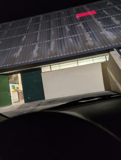 The image shows a view from the driver's seat of a car looking out at a building in Cestas, France (latitude: 44.76, longitude: -0.684). The building has a corrugated metal roof with a red rectangular object on the roof. There is a row of windows beneath the roof and the wall is white. There is a green door and a green wall on the left side of the image.  The ground is paved with concrete. There is a small object in the center of the ground.  The windshield is dark and the interior of the car is not visible.  The car is facing the building. The image is taken at night.