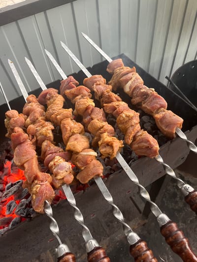 A close-up, elevated shot shows multiple rows of raw meat skewers being grilled over hot coals in an outdoor setting. The skewers, made of metal with twisted handles, are laden with cubed, marinated meat that appears to be pork or lamb. The meat is seasoned with spices that are visible on the surface. The skewers are arranged in a grill with a dark metal basin and sit above glowing red-orange coals, indicating they are actively cooking. In the background, a grey, corrugated metal wall is visible, suggesting an outdoor kitchen or patio area. The overall scene conveys the preparation of a barbecue meal. The image is likely taken in Байшуақ, Kazakhstan, given the prompt's specification, however, no visual elements within the image directly confirm this location.
