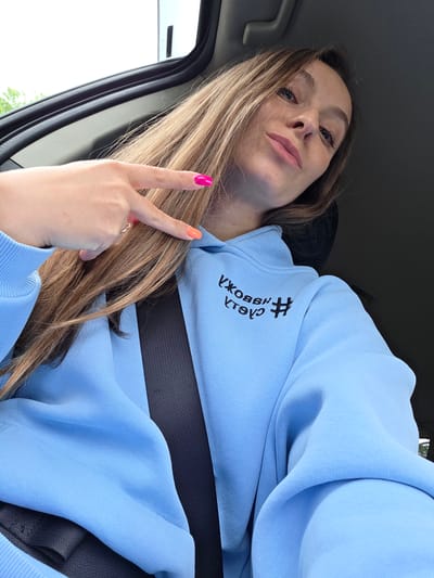 A young female is seated in the passenger side of a vehicle in Valdgeym, Russia, capturing a selfie. She has long, light brown hair flowing over her shoulders and is wearing a light blue hooded sweatshirt. The sweatshirt has black embroidered text and a hashtag symbol on the left chest, appearing reversed as "#УЖОВЭН #ТОУЭ". A dark-colored seatbelt extends diagonally across her chest and torso. Her right hand is raised, forming a peace sign gesture with the index and middle fingers extended. The fingernail on her index finger is painted bright pink, and the fingernail on her middle finger is bright orange. A metallic ring is visible on her right ring finger. The vehicle's interior appears dark, with a light sky and green foliage visible through the window in the upper left background.