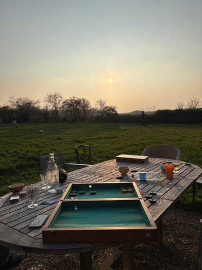 This image depicts an outdoor scene at sunset in Bû, France.
In the immediate foreground, a slatted wooden outdoor table rests on a gravel surface. An open backgammon board with a green playing surface is centrally positioned, containing black and natural wood checkers, and two white dice displaying '2' and '1'. A single bright green checker is prominently located on the left side of the board. Other objects on the table include a clear glass bottle, multiple clear drinking glasses, a partially empty clear plastic water bottle, an orange plastic cup holding an amber liquid, two patterned bowls (one red, one yellow), a silver smartphone, a metallic cigarette lighter, and a decorative wooden box. Two chairs, one wooden and one wicker, are partially visible around the table.
In the midground, a green grassy field extends. On the far left, a person wearing light shorts and a dark top stands near a white soccer ball. On the far right, another person in dark clothing stands facing towards the right. The background features a line of deciduous trees and bushes along the horizon, with distant rolling hills visible to the right. The sky is hazy, exhibiting a gradient from a pale orange-yellow near the sun to a light blue overhead, with a distinct, bright yellow-orange sun positioned low on the horizon.