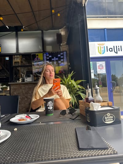 A blonde woman sits at a table outdoors, holding an orange smartphone in her hands. She is wearing a cream-colored t-shirt. In front of her is a black and green coffee cup with the logo "TO GO" and a white plate with a cookie and wrapper on it. To her right, a wooden crate holds two glass bottles, one with dark liquid and the other with brown powder. A black tissue box with the "Estrella Galicia" logo is beside the crate. Sunglasses rest on the table. The background shows a street with blue awnings and a sign that reads "la Vila." The setting appears to be a cafe or restaurant in the city of La Vila Joiosa, Spain.