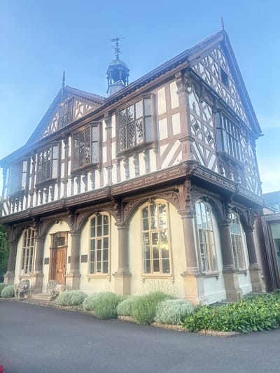 The image shows a detailed, three-story Tudor-style building. The facade is characterized by its distinctive half-timbered construction, with dark wooden beams set against a light-colored plaster. The upper floors feature numerous diamond-paned windows, many of which are in bay window configurations. Intricate wooden carvings adorn the eaves and decorative elements are present along the facade. The ground floor has a pale yellow stucco finish and is punctuated by arched windows with similar diamond panes. A wooden door is centrally located on the ground floor. Flanking the entrance steps are two stone lion statues. In front of the building, there is a paved pathway or road and a garden with low, bushy plants and some taller greenery. The sky is clear and blue, suggesting daylight. The building appears to be situated in a well-maintained outdoor setting. The architectural style and potential historical significance suggest this could be a public building or a notable residence. Based on architectural clues, it is likely the Grangefield Grange in Leominster, United Kingdom, which is known for its historical Tudor architecture.
