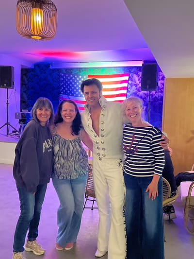 A group of four people poses for a photo in an indoor setting. From top to bottom, a woman in a navy and white striped shirt with an anchor emblem stands next to a man dressed in a white Elvis-style jumpsuit. Below them is a woman wearing a grey and white top and the bottom-most person is wearing a navy sweater. All four are wearing blue jeans.

Behind the group is an American flag and some leafy wallpaper with blue lighting. A black speaker sits on a stand in the lower left of the frame. A hanging light fixture is visible on the far left of the frame. The scene is lit with colorful ambient lighting.

Given the lack of landmarks and specific architectural details, it's not possible to confirm the precise location in Grimaud, France.