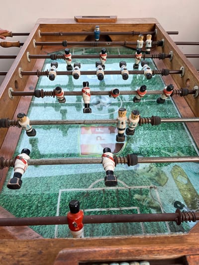 A slightly high-angle, indoor close-up shows a vintage foosball table in a wooden frame. The table features metal rods with small, multi-colored plastic players attached. The players are primarily white with black ends and some red or blue markings. The playing surface is visible in the background, depicting a green soccer field with faint logos and text. The rods extend through holes in the wooden frame, and some are equipped with springs and metal fittings. The overall condition of the table suggests age and use. It is not confirmed that the photo was taken in Pacarán, Peru.