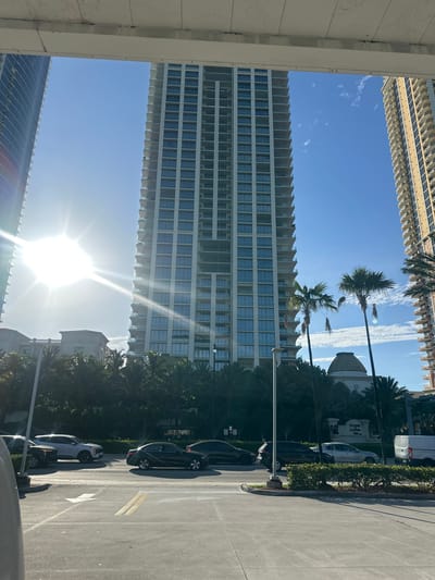The image captures a sun-drenched street scene in Sunny Isles Beach, Florida. Dominating the frame are several tall, modern residential buildings, with a particularly prominent, light-colored skyscraper in the center. To its left and right are other high-rise structures, one with a dark, curved facade and another with a beige, stucco-like exterior. Lush palm trees and other tropical foliage line the street in front of the buildings. Several cars are visible on the road, including a white SUV, a dark sedan, and a black SUV. The sky is a clear blue, with a few wispy clouds. A sign for "THE RITZ-CARLTON" is visible in the background, suggesting a high-end location. The sun is bright, casting strong sunlight and lens flares across the scene. The image appears to be taken from a slightly elevated vantage point, possibly from a parking garage or another building.