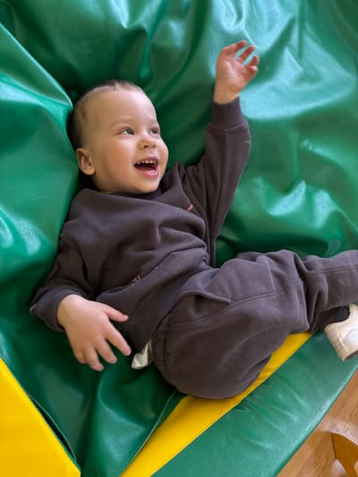 A child, wearing a dark-colored sweatshirt and pants, lies on a green and yellow mat. The child is on their back with their legs raised in the air and their hands outstretched. The child's mouth is open in a smile or laugh. The location is not possible to be determined based on visual information.