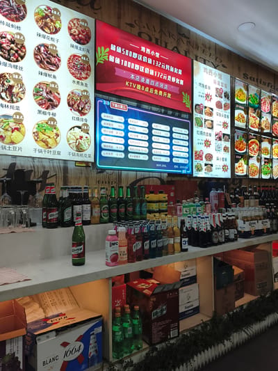 The image depicts the interior of a restaurant, likely in Jixi, China, featuring a digital menu board displaying food items and prices, alongside various beverages on shelves. The menu board, illuminated with bright colors, shows pictures of dishes such as "Spicy Shrimp", "Stir-fried Beef", and "Spicy Cuttlefish". Below the menu board, a shelf is stocked with an assortment of bottled drinks, including beer bottles from brands like "TSINGTAO" and "CORONA", as well as some unidentified bottled beverages in various colors. Further down, stacked boxes of beverages, including what appears to be "1664 BLANC" beer, are visible. The overall ambiance suggests a casual dining establishment, possibly a fast-food or a casual restaurant, focusing on Chinese cuisine and beverages. There are no people visible in the frame.