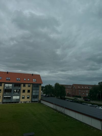 The image shows an exterior view of an apartment building complex under a cloudy sky in Roskilde, Denmark. The foreground features a well-maintained grassy lawn with a dark-colored bench. Beyond the lawn, a long, white structure with a dark, corrugated roof runs horizontally across the frame, likely a garage or storage area, with a line of parked cars visible beyond it.
To the left of the frame, a multi-story apartment building with a red tiled roof and yellow brick facade is prominent. It has numerous windows, some with balconies. To the right of this building, a row of similar apartment buildings with red tiled roofs and brick facades extends into the background. Mature trees line the area between the buildings and the street. The sky is overcast with dense, grey clouds, suggesting a possibly gloomy or impending weather event. No people are visible in the scene.