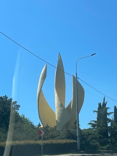 An abstract, cream-colored sculpture resembling three large, overlapping petals or flames stands against a clear blue sky. The sculpture is tall and its smooth, curved surfaces are illuminated by sunlight. To the right of the sculpture, a modern street lamp with a single light source extends upwards. A thin black wire stretches diagonally across the upper portion of the image. In the middle ground, dense green foliage, including trees and hedges, surrounds the base of the sculpture. A small, red and white directional road sign with a chevron pointing left is visible in the lower-left portion of the foliage. The foreground shows a road and a reflective surface, possibly a car window, creating a slight glare on the left side of the image. The scene is set outdoors, likely in a public area within the city of Novomikhaylovsky, Russia, given the style of the sculpture and street lamp which are consistent with public art installations found in Russian cities. There are no people visible in the image.