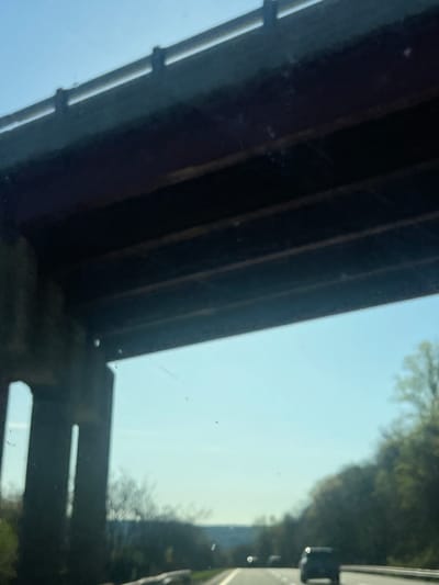 The image shows a view from under a bridge, possibly a road overpass or a railroad bridge, located in the Town of Fishkill, United States of America. The bridge structure is dark and appears to be made of steel or concrete. On the right side of the image, there is a view of the sky, trees, and a road with vehicles. The photograph is taken from a low angle.