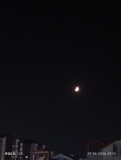 The image shows a nighttime view of a crescent moon in a dark sky over the city of Körfez, Turkey. In the lower portion of the image, the outlines of buildings and rooftops are visible. The text "POCO C65" is overlaid on the bottom left, and "20.06.2026 23:15" is overlaid on the bottom right, indicating the device used and the date and time the photo was taken. There are no people visible in the image. The overall scene is dark and illuminated only by the faint light of the moon and some artificial lights from the buildings.