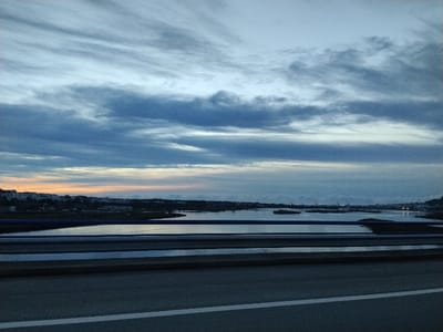 A low-angle view shows a body of water with small islands, reflecting a cloudy sky tinged with orange near the horizon. The land is visible on the horizon. A portion of a bridge with a gray surface and white lane marking is in the foreground. A blue railing separates the road from the view. The sky has layers of gray and blue clouds.