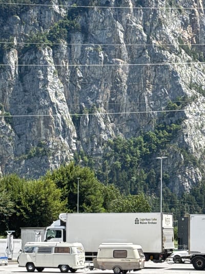 A bright, high-angle outdoor medium shot captures a parking area in front of a sheer, grey, rocky mountain face in Schattdorf, Switzerland. The mountain is partially covered with green trees and appears in the background. In the foreground, three trucks are parked. The first truck on the left is white with a black cab and is mostly obscured by a white trailer. The second truck, in the center, is also white with a black cab and a white trailer. The trailer has the logo "Lagerhaus Lohn Maison Virchaux" printed on it. The third truck on the right is partially visible and appears to be a white trailer.

Between the first and second trucks, a vintage white camper van is parked, facing left. To the left of the camper van, there are two white charging stations. In front of the camper van, a very small, light blue vehicle is parked. A light brown and cream colored caravan is parked behind the camper van. The scene is illuminated by bright sunlight, casting shadows and highlighting the textures of the rock face.