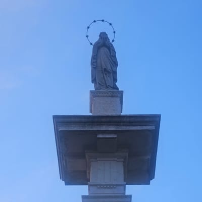 The image shows a statue of the Virgin Mary on top of a stone monument, set against a clear blue sky. The statue depicts the Virgin Mary with her hands clasped in prayer and a halo of stars above her head. The monument is made of stone and has multiple tiers and decorative elements. The lower part of the monument is a square pedestal with some relief carvings. Above the pedestal is a wider rectangular section with more elaborate architectural details. The statue is positioned on the topmost level of the monument. The scene is likely outdoors, given the sky.

This monument is located in Calcinate, Italy, specifically in front of the Church of San Pantaleone. It is known as the "Madonnina del Ponte" (Little Madonna of the Bridge) and was erected in honor of the Virgin Mary and as a symbol of protection for the bridge that was once located nearby.