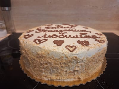 A close-up, eye-level shot shows a round cake with white frosting and chocolate decorations. The cake is covered on the sides with what appears to be crushed nuts or crumbs. The top of the cake features piped chocolate writing and several chocolate hearts. The writing is in Russian and appears to read "С днем рождения, любимая" which translates to "Happy birthday, my love." The hearts are arranged around the text. The cake rests on a black, reflective surface, possibly a stovetop. In the background, a glass salt or pepper grinder is visible, along with a blurred kitchen counter and backsplash. The lighting suggests it is daytime indoors. The setting appears to be a home kitchen in Mińsk Mazowiecki, Poland.