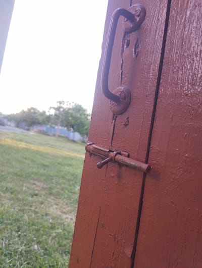 An exterior, eye-level, medium shot captures a weathered, red-painted wooden door. A curved metal handle is visible on the upper portion of the door, attached with two circular fixtures. Below the handle, a metal bolt latch is secured to the door's surface, with the bolt extended horizontally. The door is divided vertically down the center by a seam between two planks. To the left of the door, a blurred background shows a grassy area with trees and a fence in the distance. The scene is illuminated by natural sunlight, suggesting it is outdoors during the day. The overall impression is one of rustic simplicity.