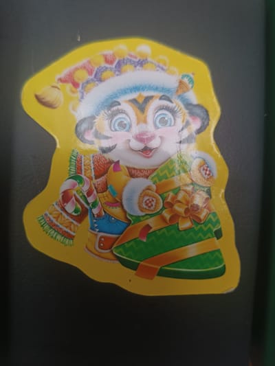 A sticker of a cartoon tiger wearing a winter hat and scarf, holding a wrapped gift, is shown against a dark background. The tiger has large blue eyes and is smiling. The gift is green with a gold ribbon and bow. The sticker is yellow and is die-cut in an irregular shape. The background is out of focus and appears to be a flat surface. The sticker is located in Skidzieĺ, Belarus.