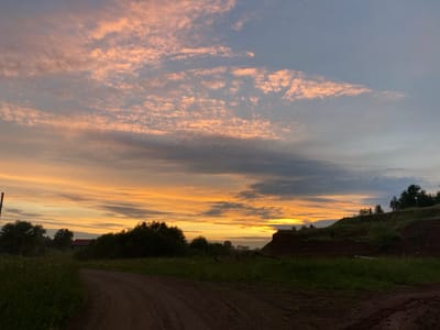 The image depicts a rural landscape in Baklushi, Russia, at what appears to be either sunrise or sunset. The sky is filled with dramatic clouds, tinged with hues of orange, pink, and gray, suggesting the golden hour. The sun is low on the horizon, casting a warm glow.
The foreground is dominated by a dirt road, curving gently to the left and leading into the scene. The road is flanked by lush green grass and vegetation, some of which is tall and wild. To the left, there is a wooden pole and some trees. Further back, a red-roofed building is visible amidst a line of dense trees.
The middle ground features a hilly terrain covered in green grass and dotted with trees. The top of the hill is lined with coniferous trees. The ground on the hill appears to be a mix of earth and vegetation, with some signs of exposed soil, possibly indicating a clearing or construction. The overall mood is serene and tranquil, capturing the beauty of a rural Russian countryside at a transitional time of day. No visible text is present in the image.