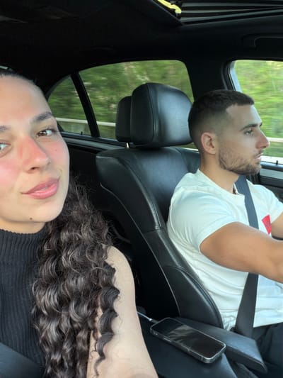 The image shows a couple in a car. The woman is in the foreground on the left, and the man is in the driver's seat on the right. Both are looking forward. The woman has dark curly hair and is wearing a black top. The man has short dark hair and is wearing a white t-shirt. They are inside a car with black leather seats and a sunroof. Outside the car, greenery is visible, suggesting they are driving through a wooded area. The location is identified as Sokolovo, Bulgaria. A phone is visible on the center console of the car, plugged into a charger.