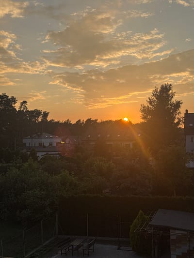 The image depicts a sunset in a suburban area, likely Marki, Poland. The sun, a bright orange orb, is partially obscured by trees and the roofs of buildings. Its rays are casting a warm, golden glow across the sky, illuminating wispy clouds that stretch across the frame. The scene is dominated by natural elements, with a dense line of trees separating the sky from the residential buildings. The architecture of the buildings appears modern, with pitched roofs and visible windows. To the right of the image, a darker section shows more dense foliage and what looks like a balcony or a fire escape attached to a building, suggesting an urban or semi-urban environment. The overall mood is peaceful and serene, characteristic of a tranquil evening. The time of day is clearly sunset due to the low angle of the sun and the warm color palette. No people are visible in the frame, and no text is present.