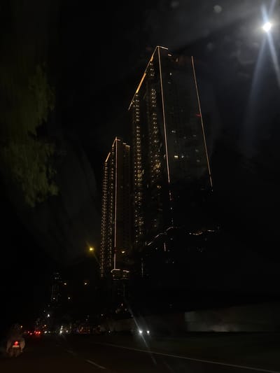 The image depicts a nighttime scene in Bencongan Indah, Indonesia. A large, dark building with multiple floors and numerous illuminated windows dominates the view. The building is outlined with linear lights along its edges. Below the building, lights suggest a parking area with rows of parked vehicles. A street with car lights runs along the right side of the frame. The sky is dark, with a bright light source visible in the upper left corner. Some blurry dark patches are visible at the bottom, likely trees or vegetation. The image is rotated 90 degrees counterclockwise.