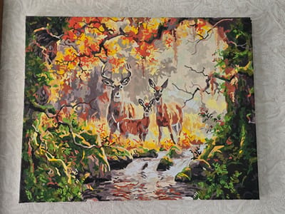 This is a painting of a forest scene with three deer by a stream. The deer are brown with white markings and have antlers. The trees are covered in autumn leaves of red, orange, and yellow. The stream is shallow and rocky, with clear water. The painting is done in a realistic style, with attention to detail in the depiction of the animals and the foliage. The painting is signed by an artist, but the signature is not legible. The painting is displayed on a white textured tablecloth. The scene does not depict any specific location in Serebriansk, Kazakhstan.