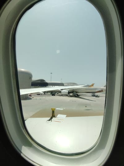 The image presents a view from an airplane window, looking out onto an airport tarmac during clear, bright daylight. The airplane window's dark gray frame and inner white trim dominate the immediate foreground, creating an oval frame for the external scene.
A large section of the white wing of the aircraft from which the photo was taken is visible in the lower foreground, extending across the frame and reflecting intense sunlight. Beyond this, another commercial airplane is parked on the tarmac. This aircraft has a white body with a notable yellow or gold-colored tailfin and what appears to be a matching stripe along its fuselage. Ground support equipment, including various vehicles and small service structures, is positioned around this parked aircraft, suggesting ongoing ground operations such as loading, unloading, or maintenance.
In the background, a modern airport terminal building with distinctive curved, gray architectural features is visible. A jet bridge extends from the terminal towards the parked airplane, and the text "FAB" is clearly visible on the side of this jet bridge. The sky is bright and clear, indicating excellent weather conditions.
The scene depicts an airplane parked at a gate, with ground crew activity around an adjacent aircraft. The bright, clear daylight and the modern airport architecture are consistent with the location context of Al Matar, United Arab Emirates.