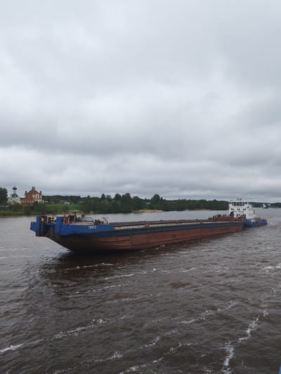 The image depicts a barge moving on a river, presumably the Volga, near Анхимово, Russia. The barge is dark brown and blue, with the number "7663" visible on its side. A tugboat is connected to the barge at its rear. The water is dark and choppy. On the riverbank to the left, a church complex with distinctive onion domes and a brick structure is visible amidst green trees and grass. The sky is overcast, creating a subdued lighting condition.