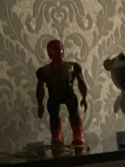 In a dimly lit room, a plastic figurine of a superhero, possibly Spider-Man due to its red and blue attire with gold accents, stands on a reflective surface. The figurine is the central subject, depicted from the waist up, with its body facing forward and head turned slightly to the left. The background features a textured wallpaper with a repeating floral or damask pattern in muted, light gray tones. To the right of the figurine, a blurry, lighter-colored object, possibly another toy or decorative item, is partially visible. The overall scene is captured in a slightly out-of-focus, grainy manner. The image does not provide any information regarding the city of Sumgayit, Azerbaijan.