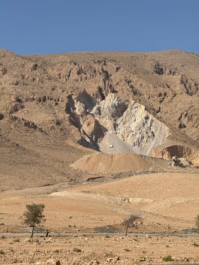 This image displays an arid, mountainous landscape located in the City of Adra labor, Syria, under a clear blue sky. The foreground consists of rocky, light-brown ground scattered with stones and sparse, low-lying vegetation. A dark asphalt road traverses horizontally across the lower portion of the frame. Two small, sparsely foliated trees stand near the road, with a dark, indistinct object positioned near the base of the left tree. The midground features barren, gently sloping terrain. The background is dominated by a rugged mountain range. A prominent excavation or quarry site is visible on the mountain face, characterized by a large exposure of light-colored rock and a substantial reddish-brown spoil heap at its base. A cluster of small, light-colored, low-rise structures is present at the mountain's base to the right of the spoil heap. No individuals or active interactions are visible in the scene.