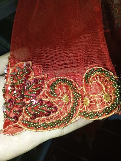 In Keo Kouang, Laos, the image presents a close-up, high-angle view of an embellished red textile draped over a human hand. The upper portion of the fabric consists of a sheer, fine-mesh red material. Below this, a scalloped border made of a denser, opaque red fabric is visible, extending diagonally across the lower half of the frame. This border is intricately adorned with gold-colored thread embroidery forming web-like and swirling patterns. Dark green/bronze seed beads are arranged in dense, curvilinear designs that follow the scalloped edge. Interspersed among these embellishments, primarily on the left side of the border, are numerous round, translucent red sequins and smaller red spherical beads, along with a few clear or iridescent beads. A human hand, displaying light skin pigmentation, is positioned beneath the textile, supporting it, with parts of the palm and fingers visible along the bottom and lower-left edge. The background behind the fabric is dark and out of focus.