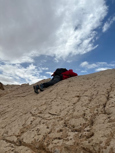 The image captures a lone individual ascending a steep, light brown, heavily fractured rock face in the Arava Tichona Regional Council, Israel. The rock surface exhibits numerous small, irregular pockmarks and extensive cracking. The person is positioned prone, oriented upwards along the incline, wearing dark-colored trousers, a bright red hooded jacket, and a black backpack. Dark green and black hiking boots are visible, with their soles in contact with the rock. The sky above transitions from clear blue with scattered white cumulus clouds on the right to a more overcast, grey cloud formation on the upper left. Distant, indistinct rocky formations are visible on the horizon in the upper left background.