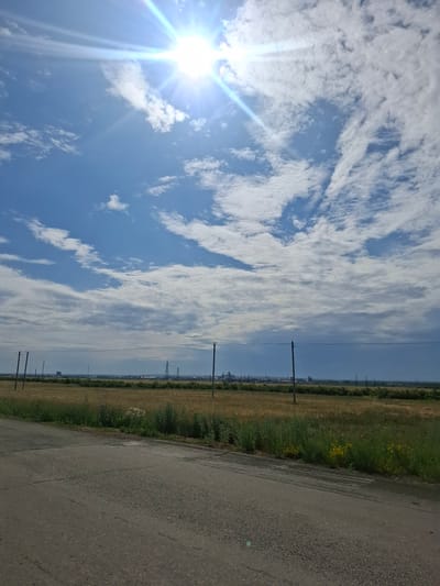 This media file captures a daytime scene of a road running alongside a field, with a bright sun and scattered clouds in a blue sky. The image is oriented vertically.
The main subjects are the natural elements: a vast expanse of sky filled with fluffy white clouds, and a sun that is prominently featured with lens flares, suggesting it is either high in the sky or the camera is pointing towards it. Below the sky, a field of dry grass with patches of green and some small yellow wildflowers stretches out. To the right of the field is a paved road, appearing somewhat weathered. Along the edge of the field, there are several utility poles with power lines, and in the far distance, some industrial structures or buildings can be faintly seen.
There are no people visible in the image. The activity taking place is simply the depiction of a landscape on a sunny day. The time of day appears to be mid-day, given the brightness of the sun and the clear blue sky, though the presence of clouds moderates the intensity. The weather seems fair and sunny. The location cues, such as the utility poles and the type of vegetation in the field, along with the mention of Atbasar, Kazakhstan, suggest a rural or semi-rural setting in a steppe-like environment. No text is visible in the image. The overall emotion conveyed is one of openness and natural beauty, with a hint of industrial presence in the background.