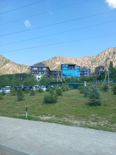 A wide shot captures a modern hotel complex nestled against a backdrop of rugged, rocky mountains under a clear blue sky in Chimgan, Uzbekistan. The hotel features contemporary architecture with large, reflective blue glass windows and solar panels adorning the rooftops. One building displays a prominent graphic of a white soccer ball set against a green background, framed by white stars. A parking lot filled with a variety of cars, including white vans, SUVs, and sedans, is situated in front of the hotel. Lush green trees and manicured shrubs surround the buildings and parking area, with a dirt road or path visible in the foreground. Power lines stretch across the top of the frame, and a single white utility pole with wires stands to the right of the hotel. The overall scene depicts a developed resort area within a natural mountain landscape.