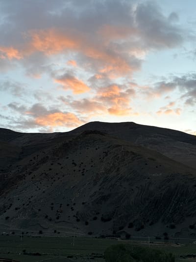 A landscape photograph depicts a mountain range against a partially cloudy sky. The sky is on the left side of the frame, featuring clouds illuminated with orange hues, suggesting either sunrise or sunset. The mountain range dominates the right side of the image, displaying dark, textured terrain. At the base of the mountain is a thin line of green, suggesting the beginning of a lower-elevation grassy area. Overhead power lines are visible in the green area, suggesting the presence of a town or infrastructure.

The image captures the contrast between the natural elements of the mountain range and the sky, with a subtle hint of human presence through the power lines, possibly near Tashi Dzom town, China.