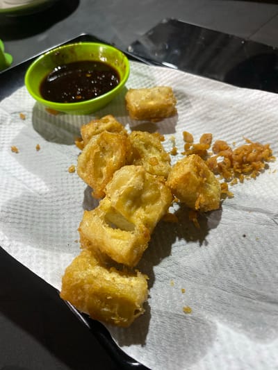 The image shows a close-up of fried tofu on a white paper towel. A small green bowl containing a dark sauce sits to the left of the tofu. The tofu pieces are irregularly shaped and golden-brown in color, with some scattered crispy fried bits nearby. The paper towel rests on a dark surface, possibly a tray or table. The location is Gampongaceh, Indonesia. There are no people visible in the image.