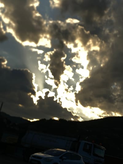 A wide shot captures a dramatic sky filled with sunbeams piercing through dark clouds, creating a striking contrast of light and shadow. Below the dramatic sky, the silhouette of a mountainous landscape is visible, with trees and hills creating a dark, textured backdrop. In the foreground, a white car is parked, facing slightly to the left. Behind the car, a large truck with a white trailer is partially visible. The truck appears to be stationary. The overall impression is a scene captured during a time of atmospheric intensity, possibly before or after a storm. The location is Prijepolje Municipality, Serbia.