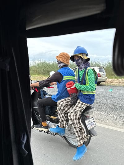 Two individuals are riding a motorcycle in Krong Ta Khmau, Cambodia. The person in front is wearing a blue hard hat, a yellow brimmed hat, and a patterned face covering. They are also wearing a blue vest over a green long-sleeved shirt with a repeating geometric pattern and a blue vest with reflective yellow trim. Their pants are checkered, and they are wearing blue rubber boots. They are holding a red thermos with a white lid. The person behind them is wearing an orange bucket hat and a blue vest over a dark long-sleeved shirt. Their pants are also checkered, and they are wearing black shoes with orange socks. The motorcycle has a license plate that reads "1JR-8867 PHNOM PENH". The background shows a road with gravel on the side, and vegetation and a light-colored SUV are visible in the distance. The sky is overcast.